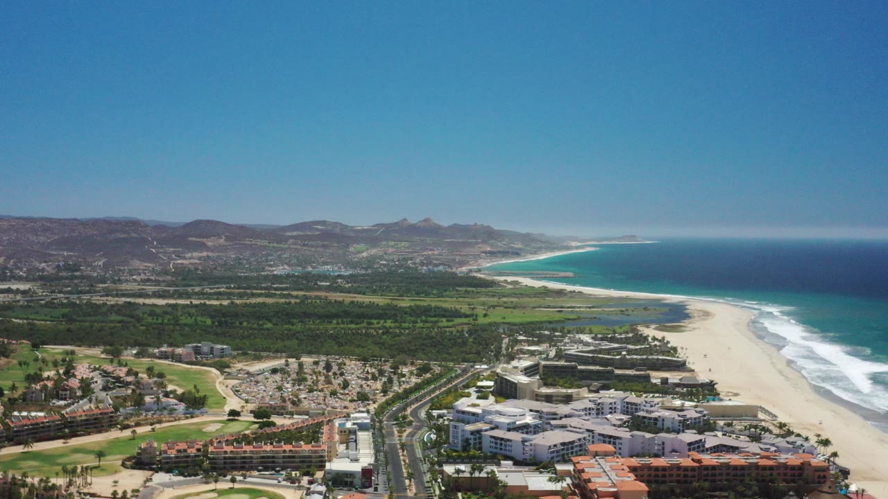 High aerial view over San Jose Del Cabo beachfront buildings, a road, green lowland, pale sand, blue ocean, distant hills, and no visible people.