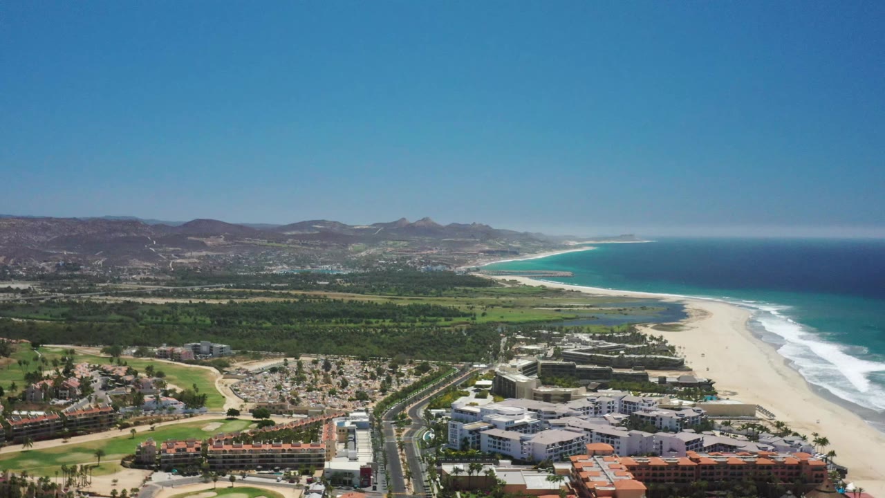 A slightly later aerial frame of San Jose Del Cabo shows the same beachfront district, curving sand, foamy surf, blue water, hills, and no visible people.