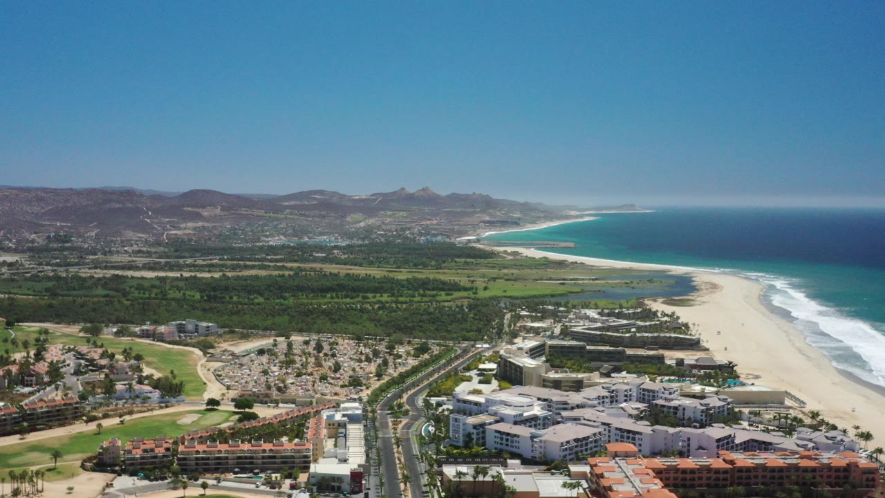 High aerial over San Jose del Cabo with beachfront buildings and a divided road in the foreground, pale beach and blue ocean to the right, inland greenery and hills beyond, with no visible people.