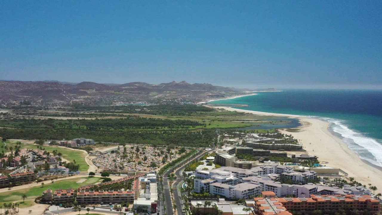 San Jose del Cabo appears from above with low-rise buildings at the bottom edge, a straight road leading toward the beach, surf breaking along the shore, and hills across the horizon, with no visible people.