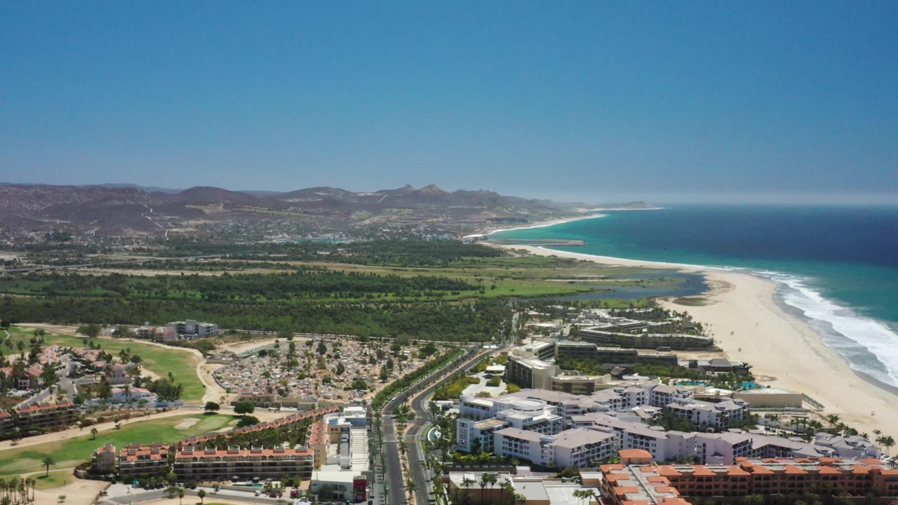 Aerial coastline with hotels, beach, ocean, green terrain, and mountains.