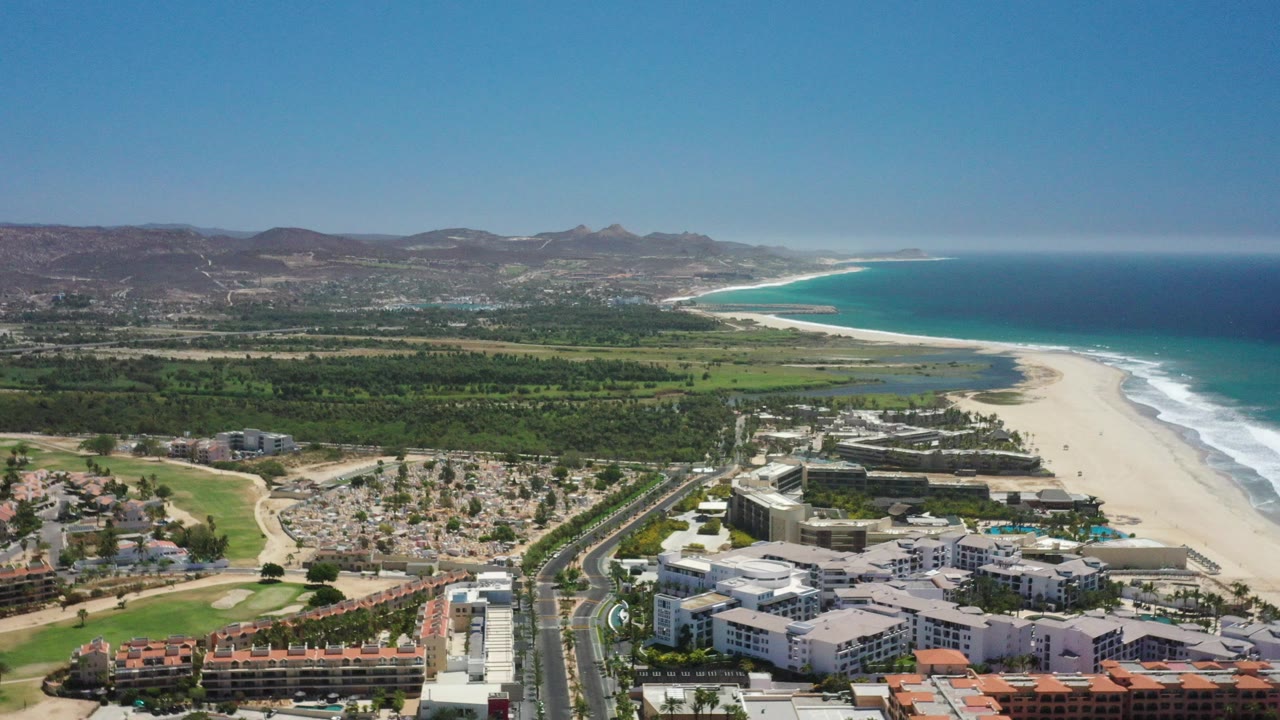 Aerial view of beachfront buildings, sandy beach, ocean, green land, and mountains.