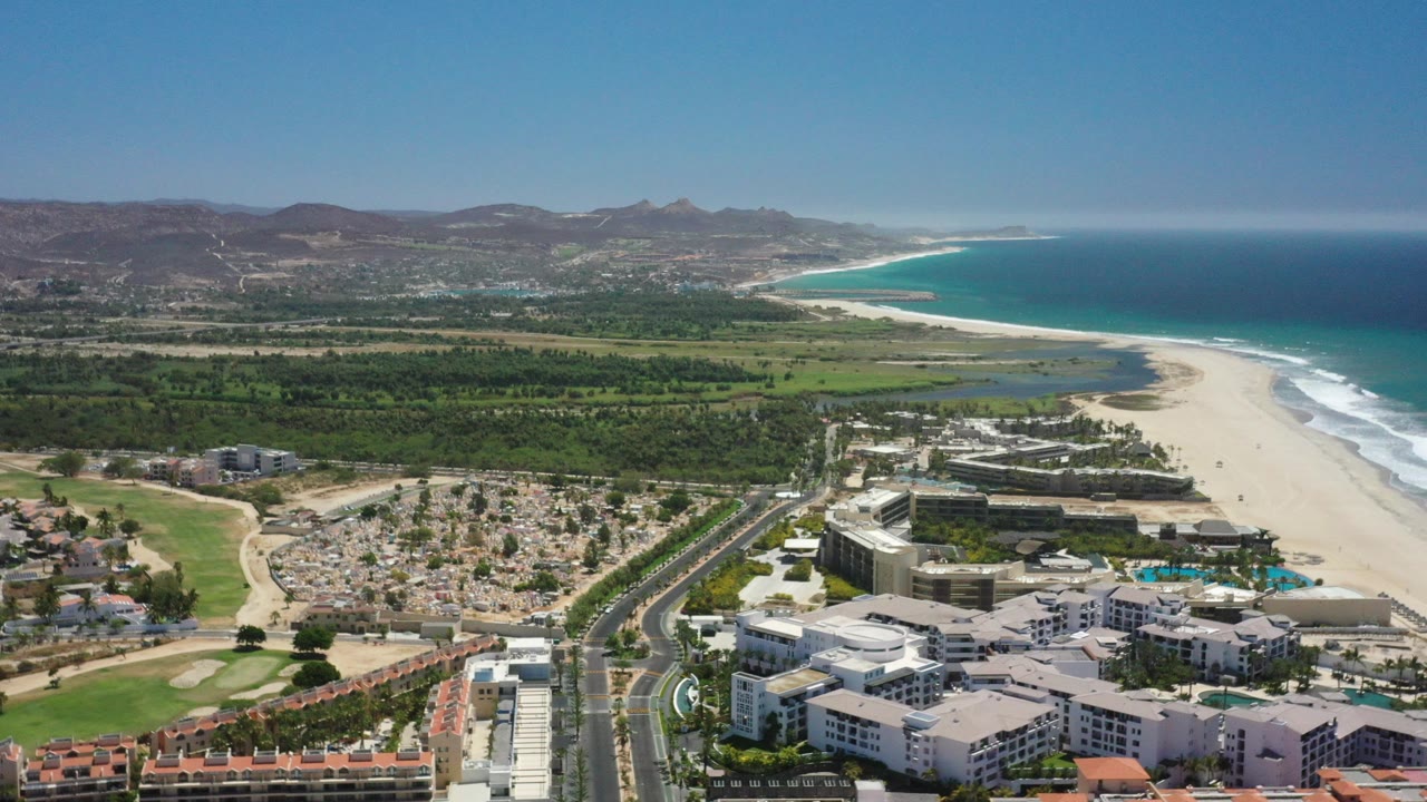 High aerial view over San Jose del Cabo, Mexico with beachfront buildings at the bottom, a road and palms through the center, pale beach and turquoise ocean to the right, distant hills, and no visible people.