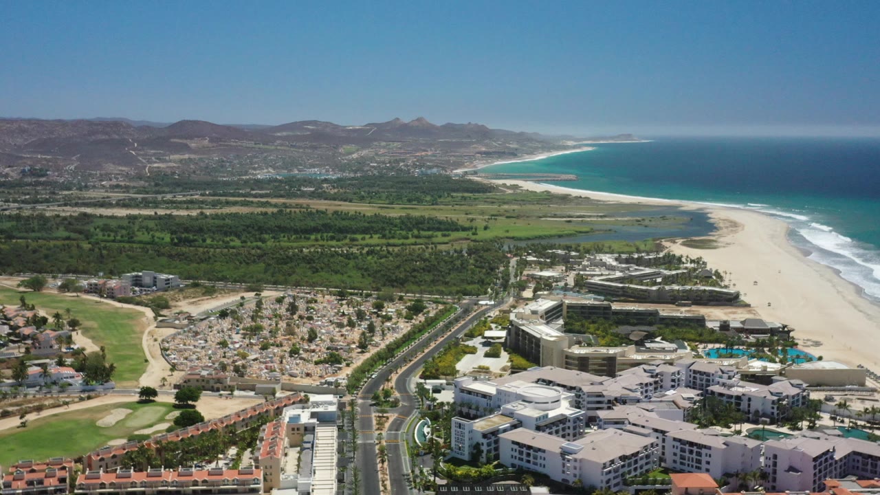 Aerial beach and ocean view above hotels, palms, inland greenery, and mountains