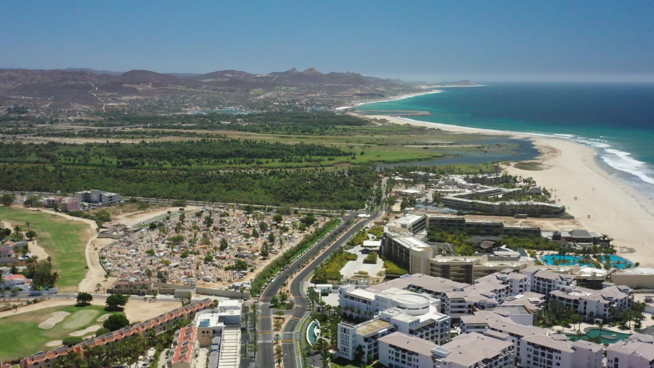 A high aerial frame over San Jose del Cabo shows resort buildings and pools in the foreground, a road and sandy cemetery area at left, green coastal vegetation, beach, ocean, distant hills, and no visible people.