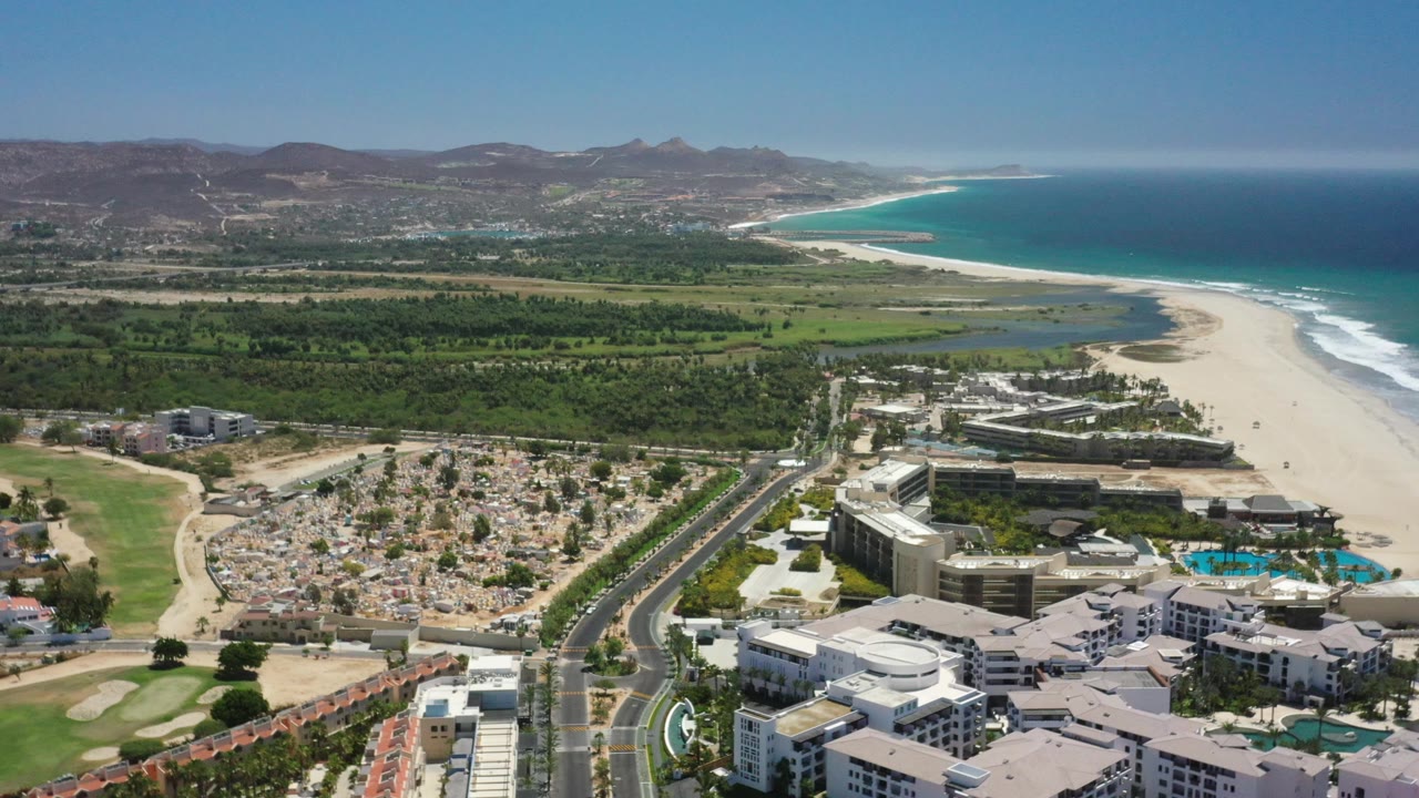 Aerial view across beachside resorts, green land, ocean, surf, and mountain backdrop.