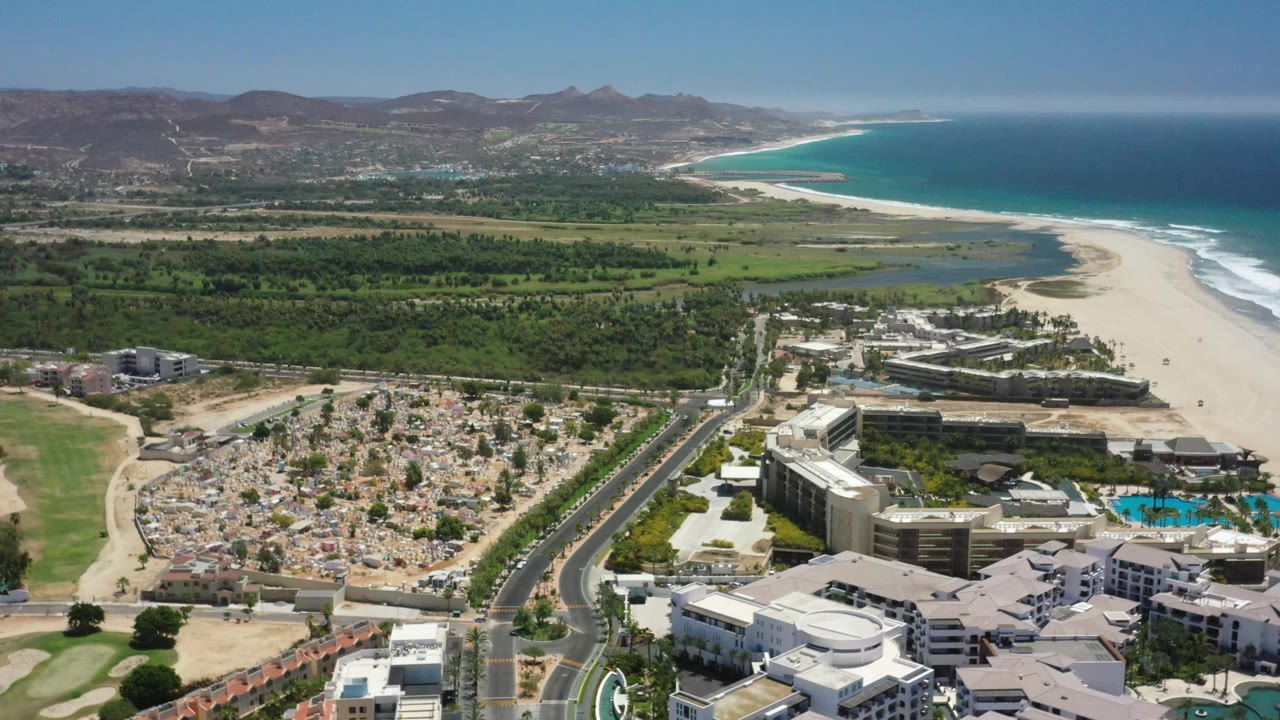 San Jose del Cabo aerial view one second into the clip, with the road and clustered buildings lower in frame, beach curving along the right side, blue water beyond it, and no visible people.