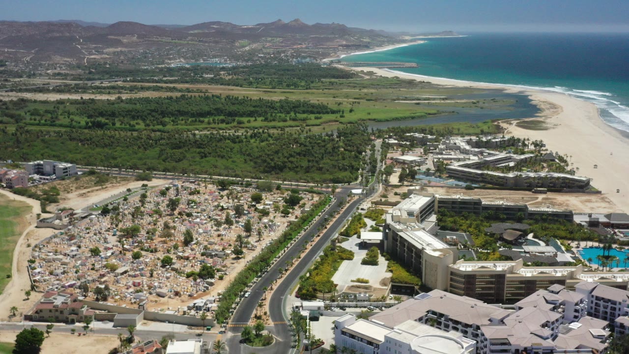 High aerial view over San Jose del Cabo, Mexico, with hotel buildings and roads in the foreground, a pale beach and blue ocean beyond, and no visible people.