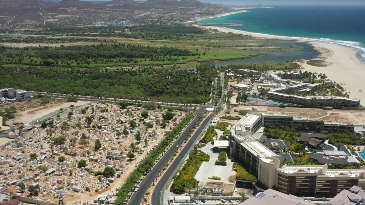 Aerial coastal city view with road, cemetery, buildings, greenery, beach, and blue sea.