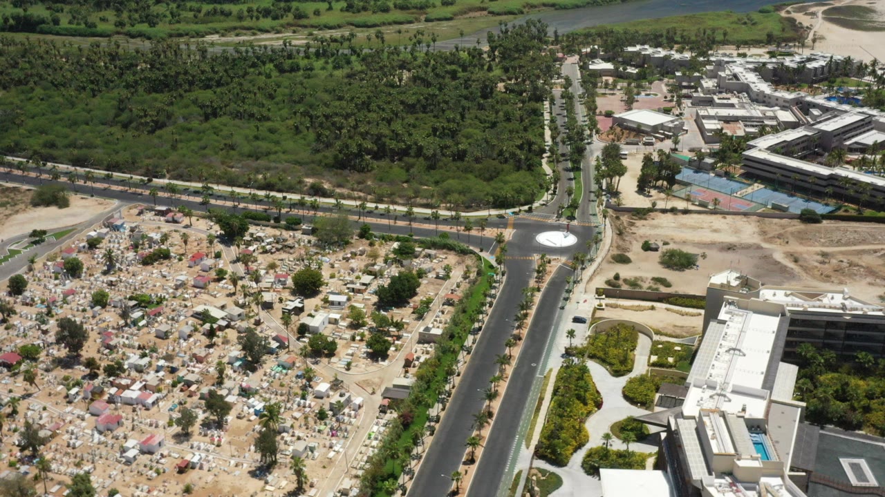 High aerial view in San Jose del Cabo, Mexico, showing a divided road, small white roundabout, dense green vegetation, nearby buildings, and a cemetery-like area with no visible people.