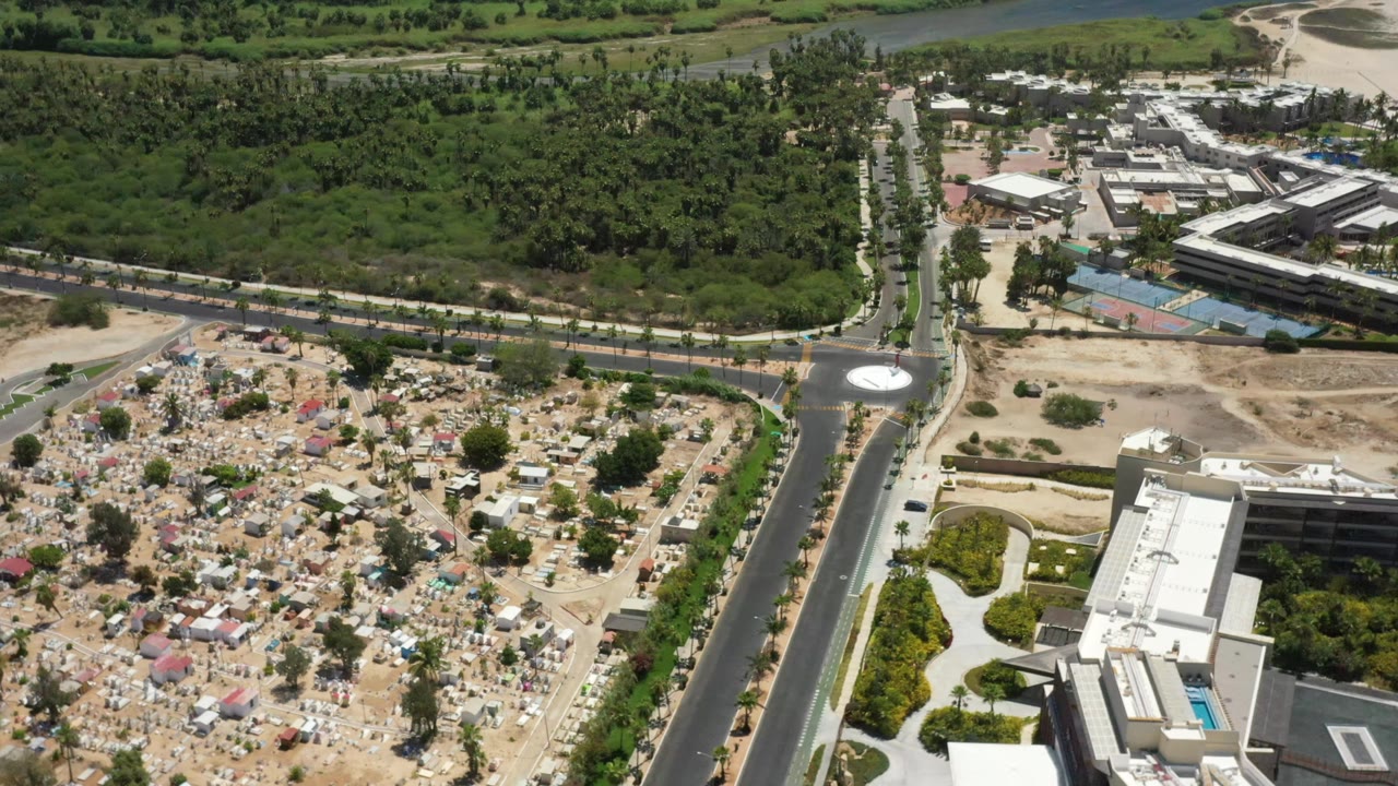Aerial frame over San Jose del Cabo with the road running vertically through the view, cemetery-like plots to the left, green vegetation at the top, and buildings to the right, with no visible people.