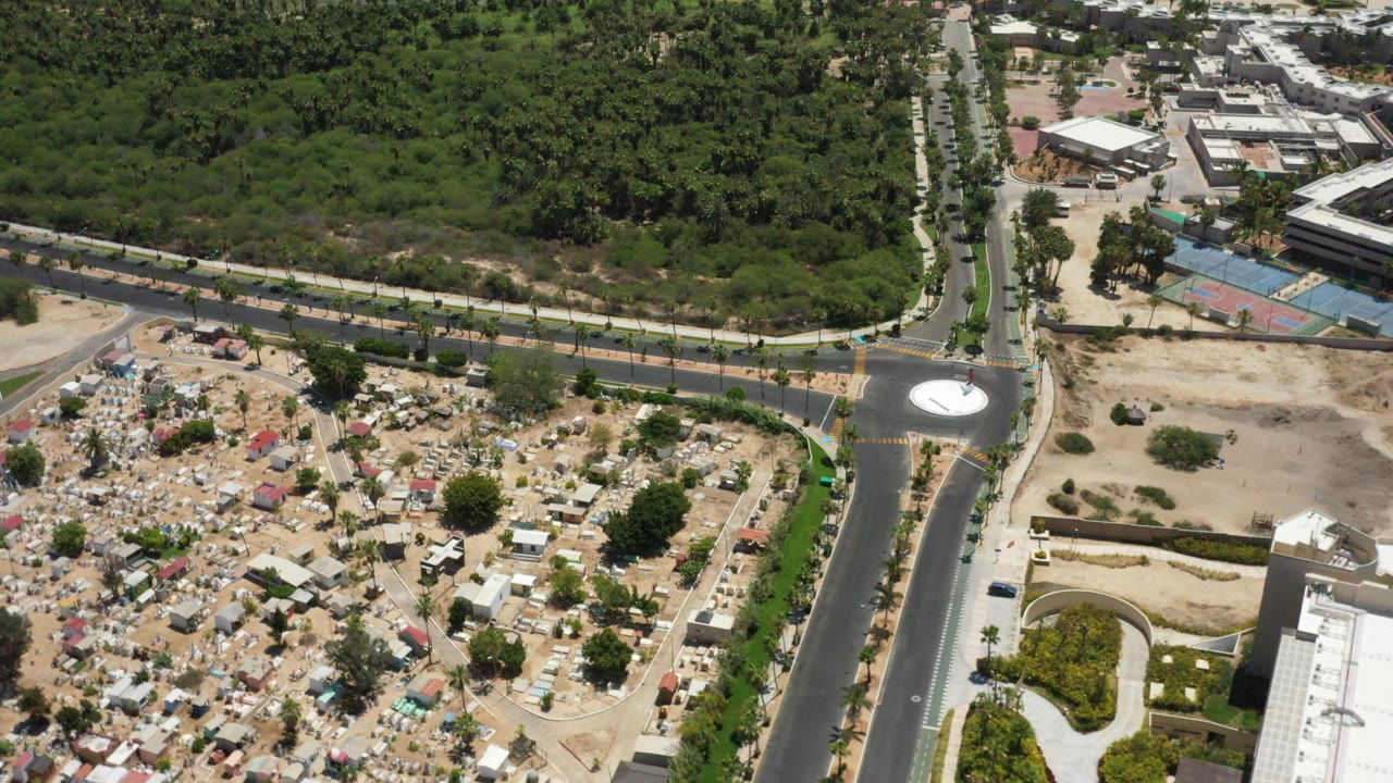 Aerial view of a road junction beside greenery, buildings, and a cemetery-like plot.