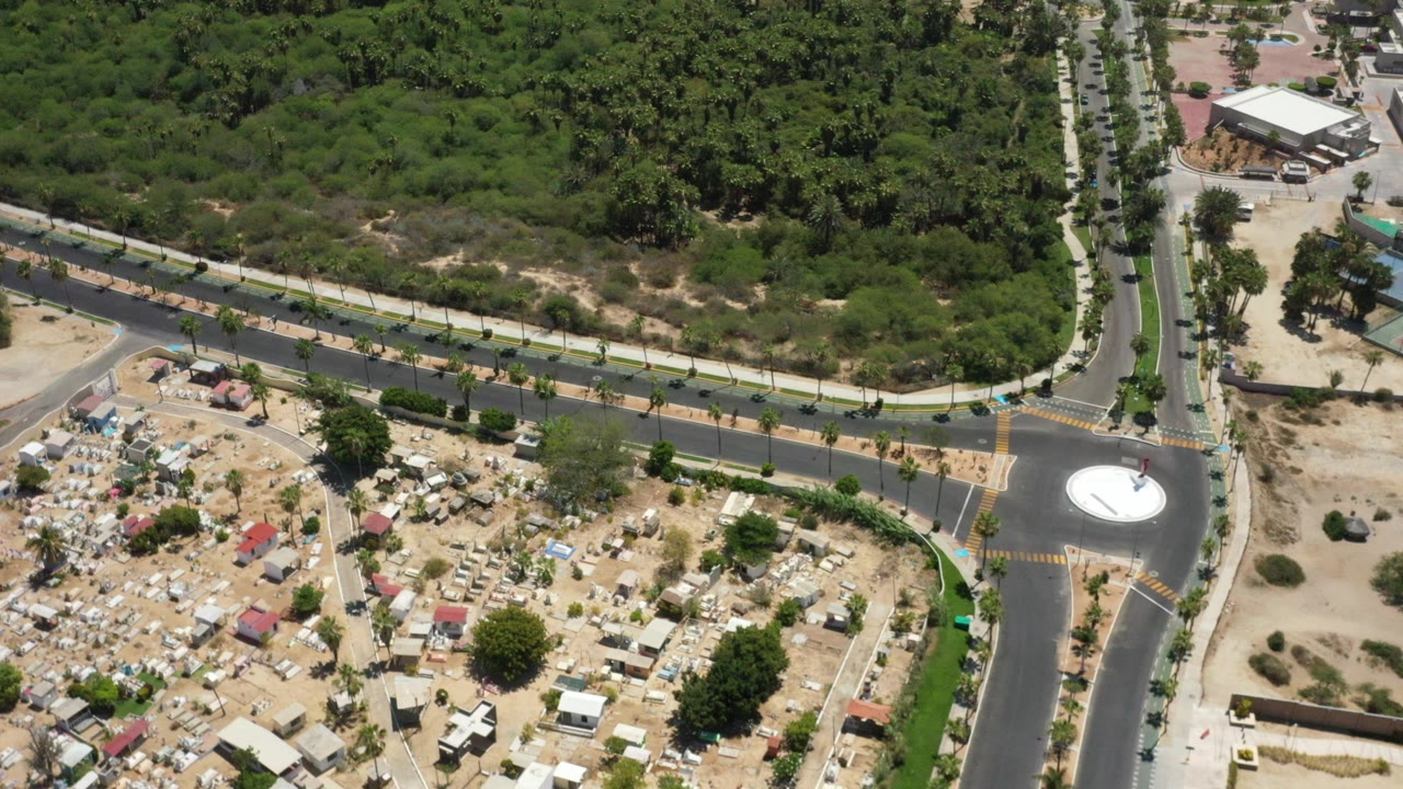 Overhead drone view in San Jose Del Cabo, Mexico showing a paved road junction beside dense green trees, sandy lots, scattered small structures, and no visible people.