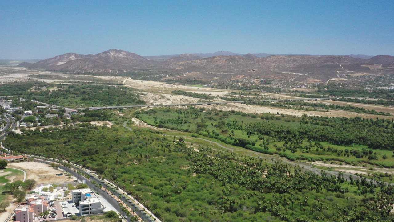 Aerial view over green valley, roads, buildings, and distant mountains under blue sky.