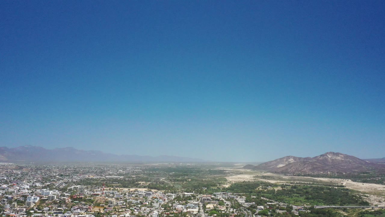 Distant aerial cityscape in San Jose del Cabo, Mexico, with white buildings low in frame, green midground, mountains on the right, haze across the far horizon, and no visible people.