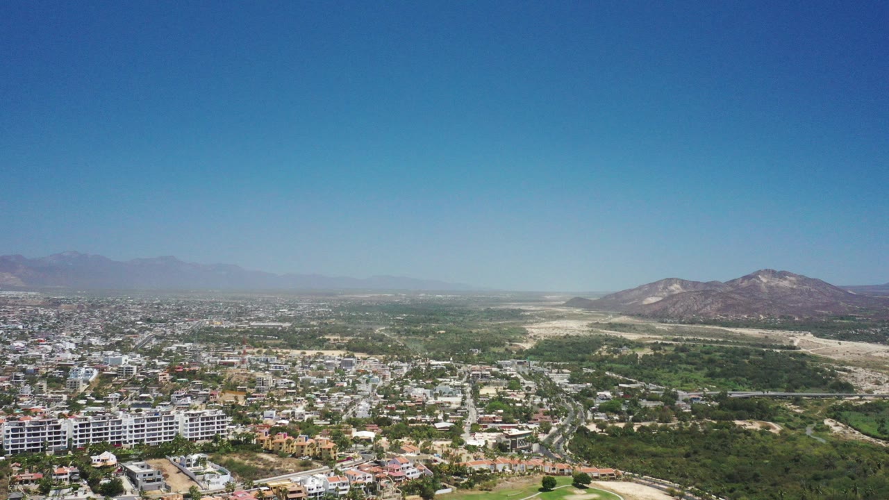 Wide aerial view over San Jose del Cabo, Mexico, showing residential buildings in the foreground, green hills through the center, distant mountains, and no visible people.