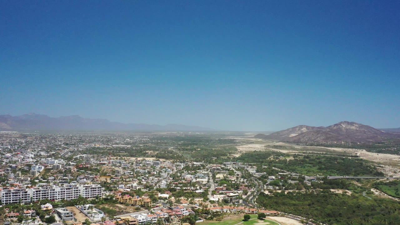 Aerial frame from San Jose del Cabo, Mexico, with white apartment blocks low in frame, a spread of homes and roads, green scrubland, distant mountains, and no visible people.