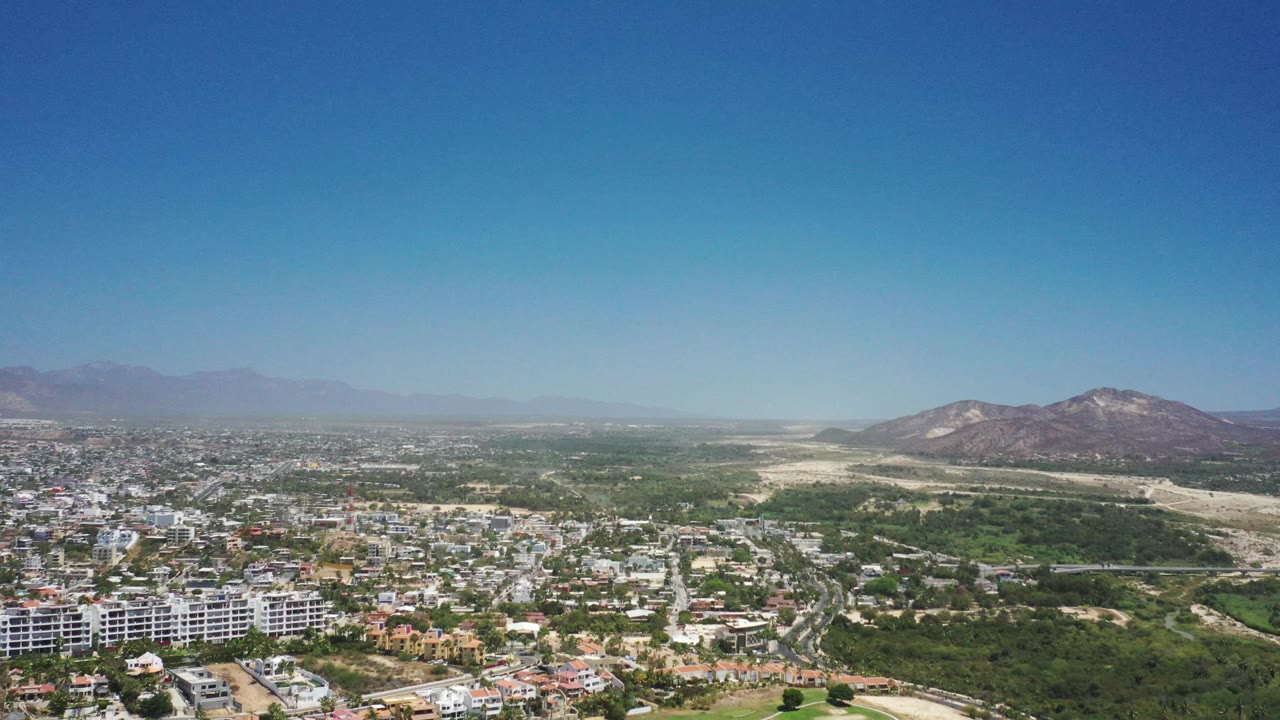 High aerial view of a town beside green hills and pale dry terrain with mountains in the distance.