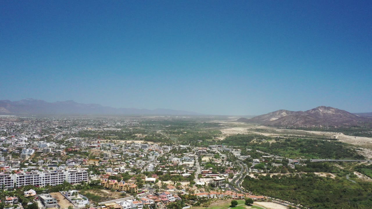 Aerial view of city blocks, green terrain, dry wash, and hills under blue sky.