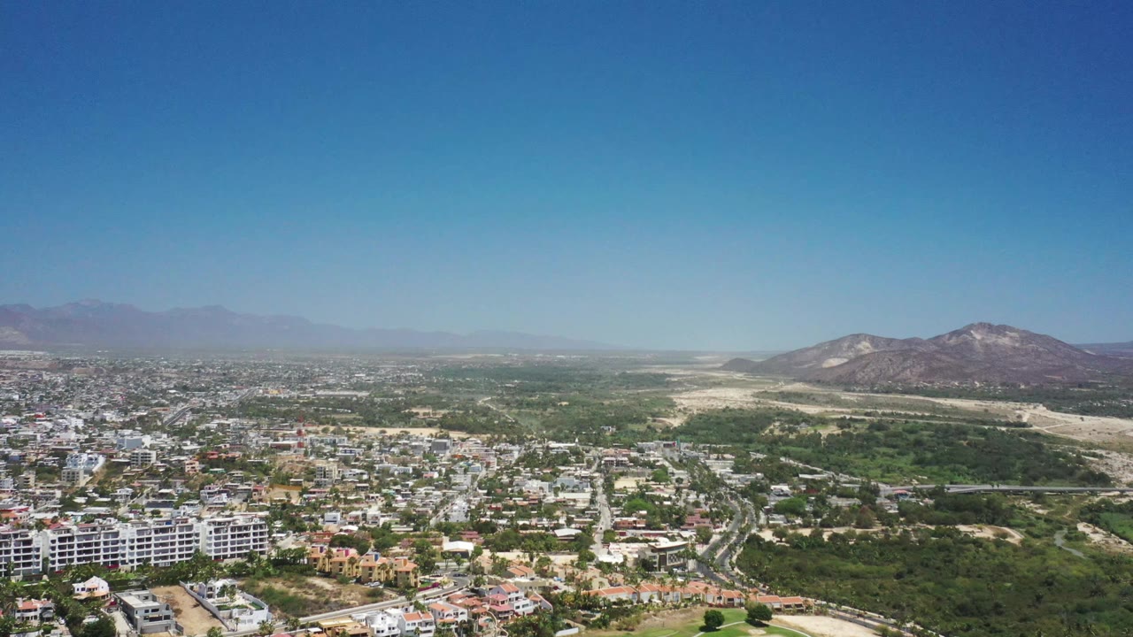 Aerial frame over San Jose del Cabo showing clustered low-rise buildings at left, a green corridor through the center, pale undeveloped ground near the hills, and no visible people.