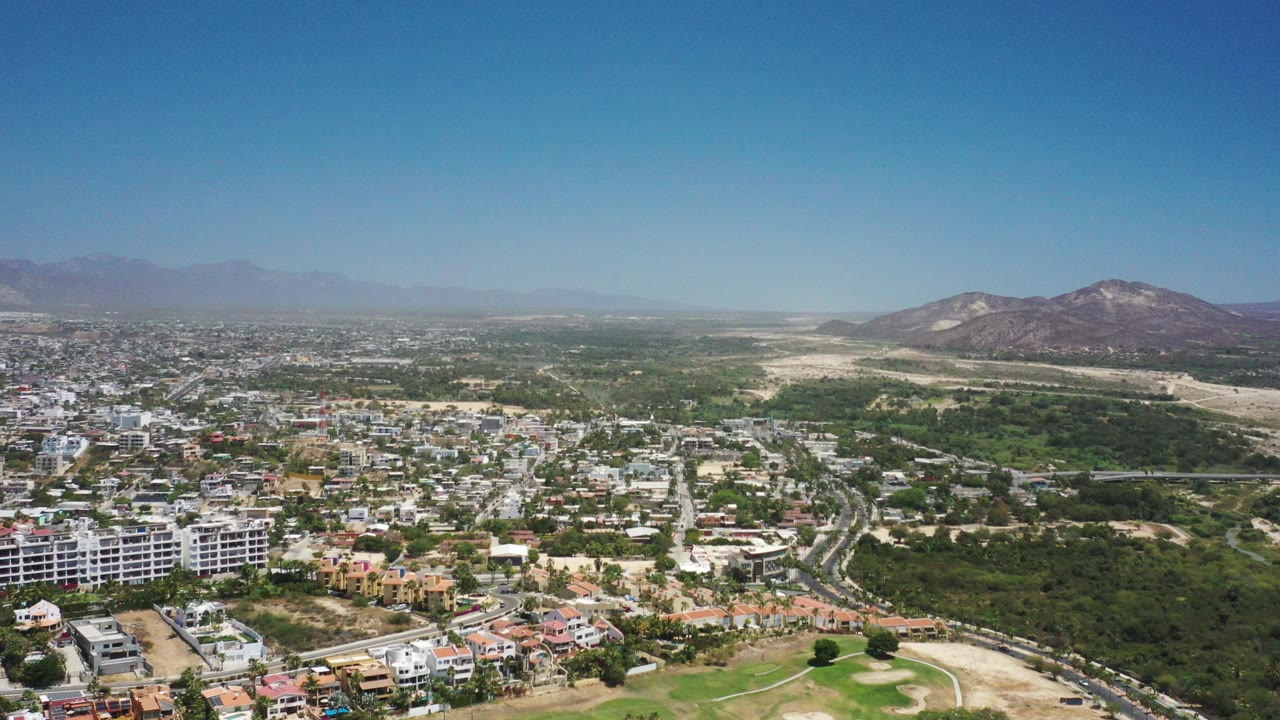 Aerial view of town buildings, green land, roads, and mountains under a clear sky.