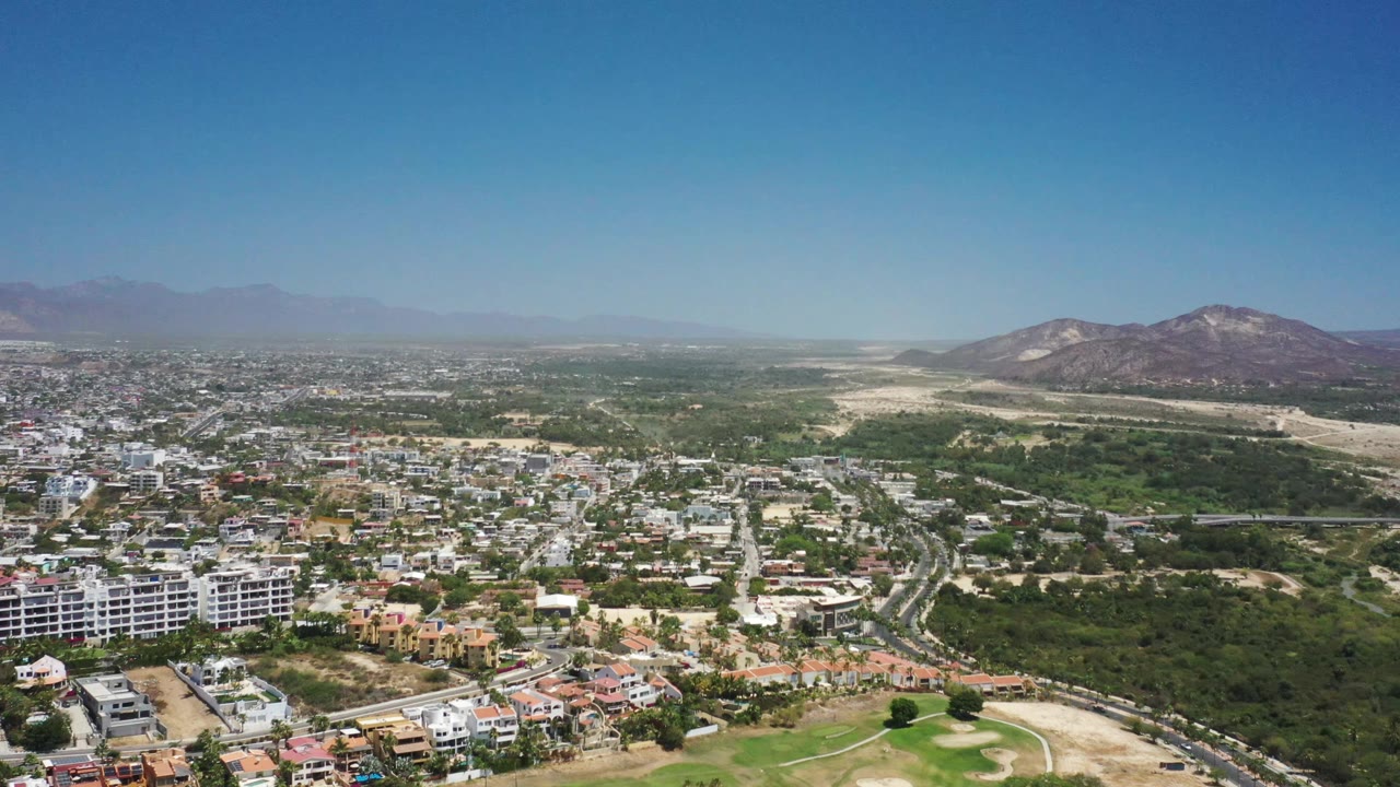 Aerial frame over San Jose del Cabo with dense town blocks at left, a broad green area in the center, dry wash beyond, mountains at right, and no visible people.