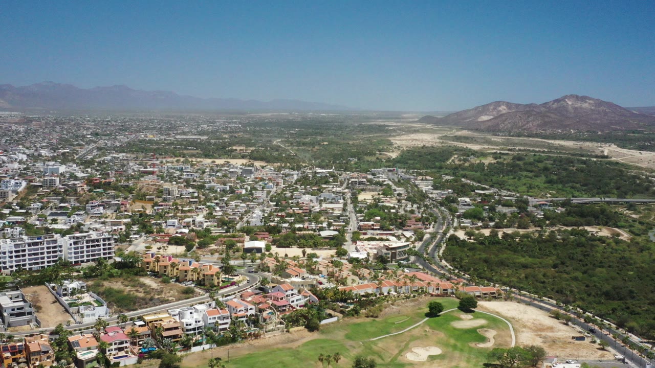 Wide aerial view over San Jose Del Cabo, Mexico, with low-rise buildings, roads, green vegetation, tan open ground, distant mountains, and no visible people.