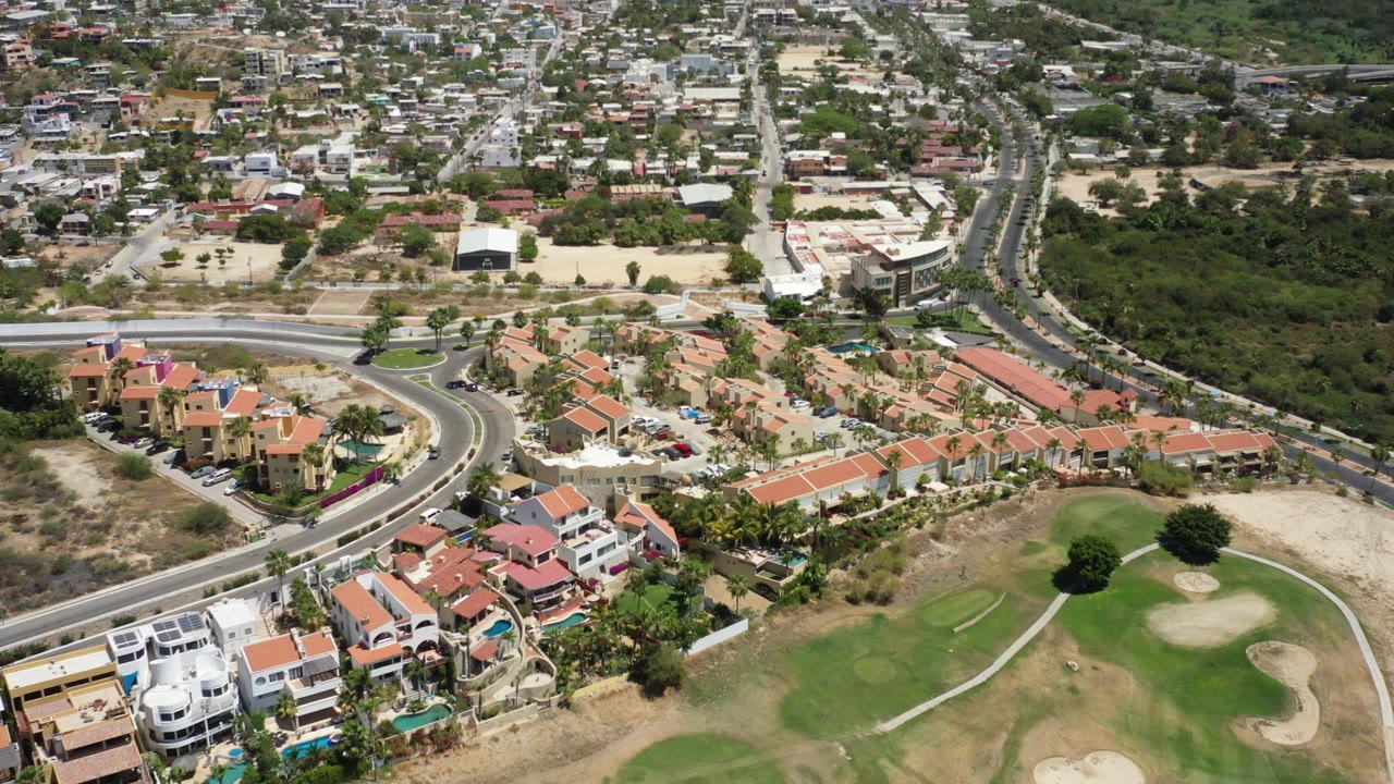 Aerial frame over San Jose del Cabo homes with red tile roofs, a curved road, green fairway, sandy bunkers, and no visible people.