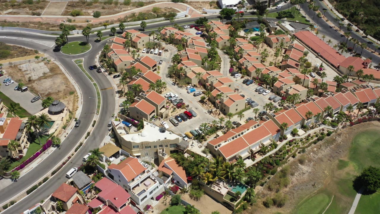 Opening aerial frame over San Jose del Cabo homes with red tile roofs, a curving road, palms, parked cars, dry lots, and no visible people.