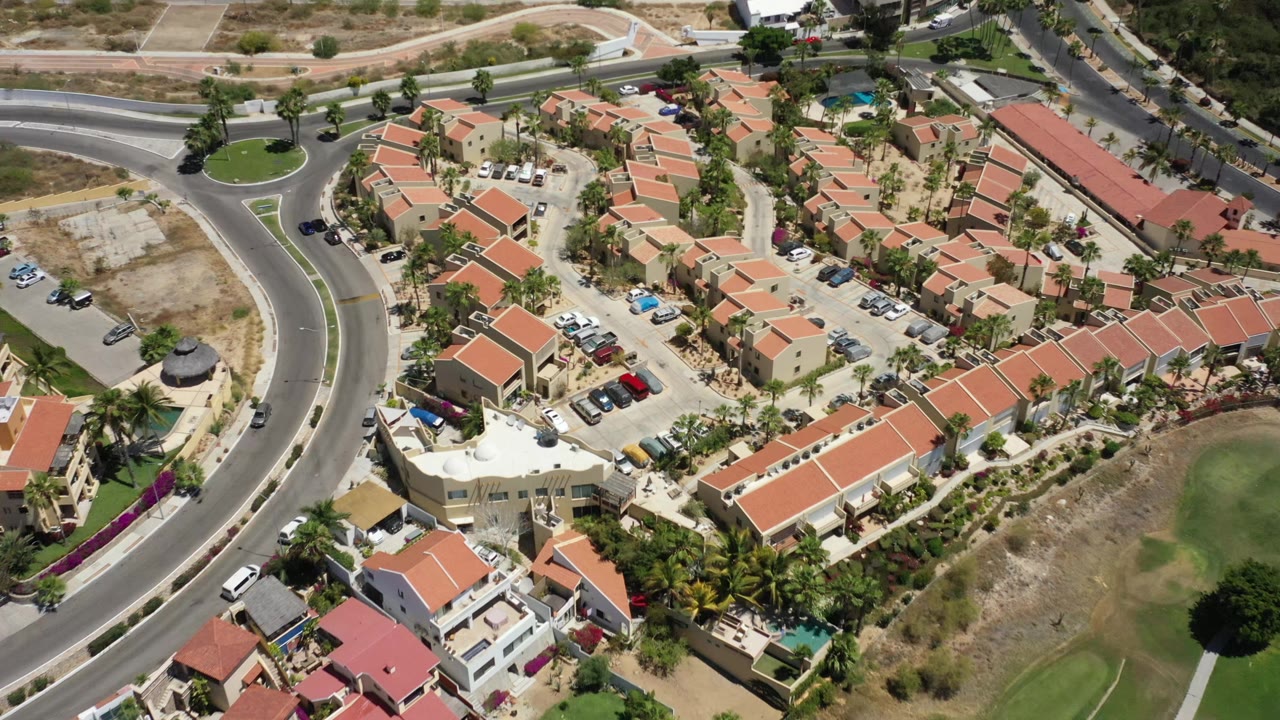 Aerial frame in San Jose del Cabo showing clustered tan houses, red roofs, parked cars, a curved road edge, dry terrain, palms, and no visible people.