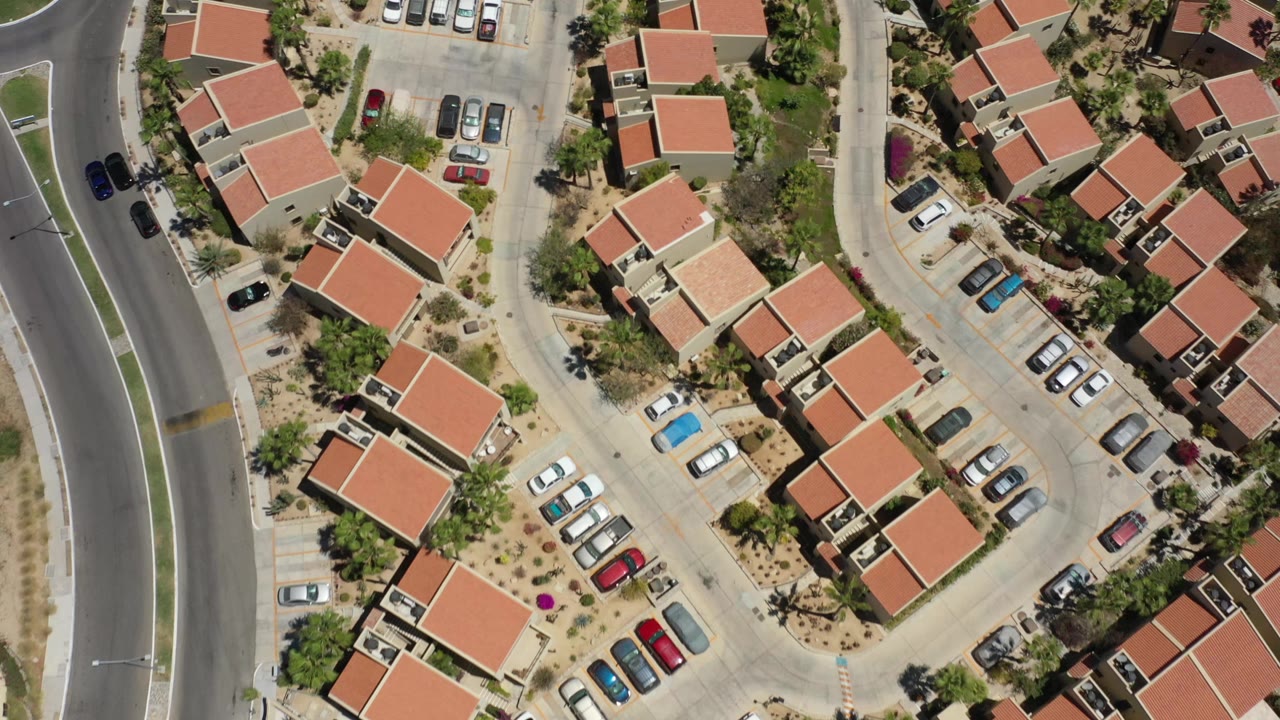 High aerial view over San Jose del Cabo residences, shared parking spaces, light-colored pavement, palms, and dry landscaping, with no visible people discernible.
