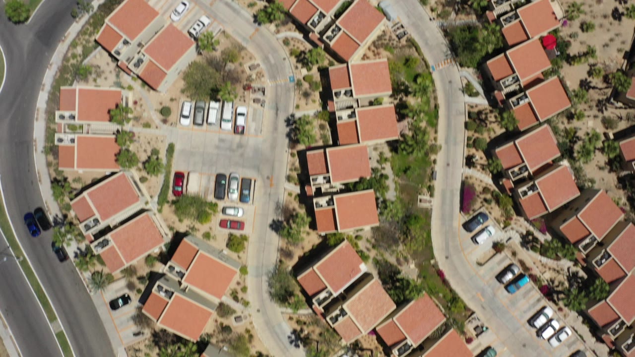 Top-down aerial view in San Jose del Cabo showing red tile-roofed homes, a curved road, parking spaces, desert landscaping, and no visible people.