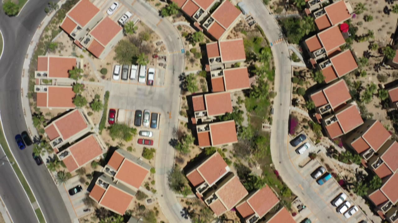 High aerial frame in San Jose del Cabo with rows of red-roofed homes, parked vehicles, pale pavement, planted yards, and no visible people.