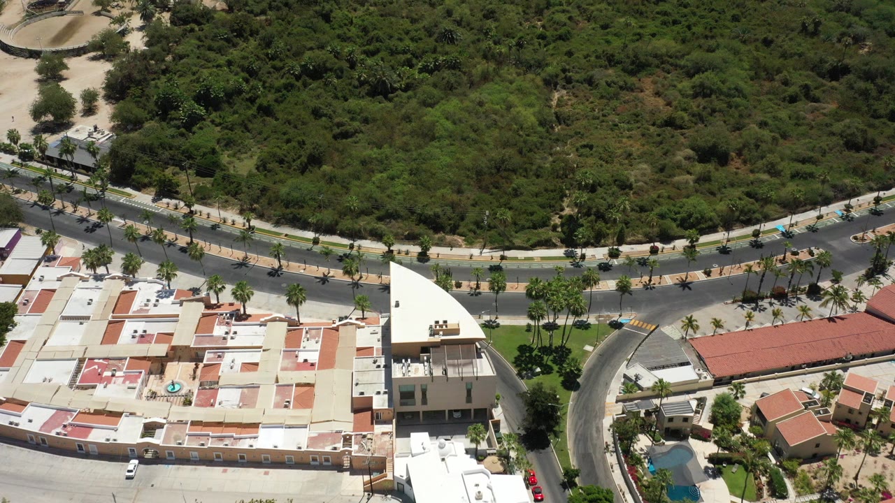 Opening aerial frame over San Jose del Cabo with pale rooftops, a curved road, palm trees, dense scrubland beyond, and no visible people.