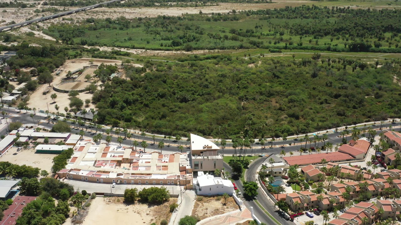 High aerial view over San Jose del Cabo with red-roof homes in the lower right, a pale building complex at lower left, curved roads, dense green trees, and no visible people.