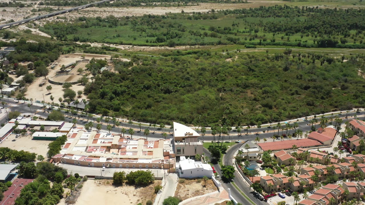 Elevated San Jose del Cabo frame with a curved road between a pale building complex and red-roof homes, dense vegetation behind them, distant fields, and no visible people.
