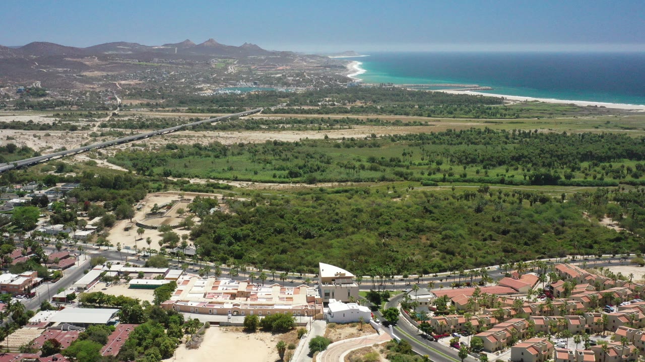 Wide aerial view over San Jose del Cabo, Mexico, with red-roofed homes in front, green lowlands and dry terrain beyond, hills on the horizon, blue ocean at right, and no visible people.
