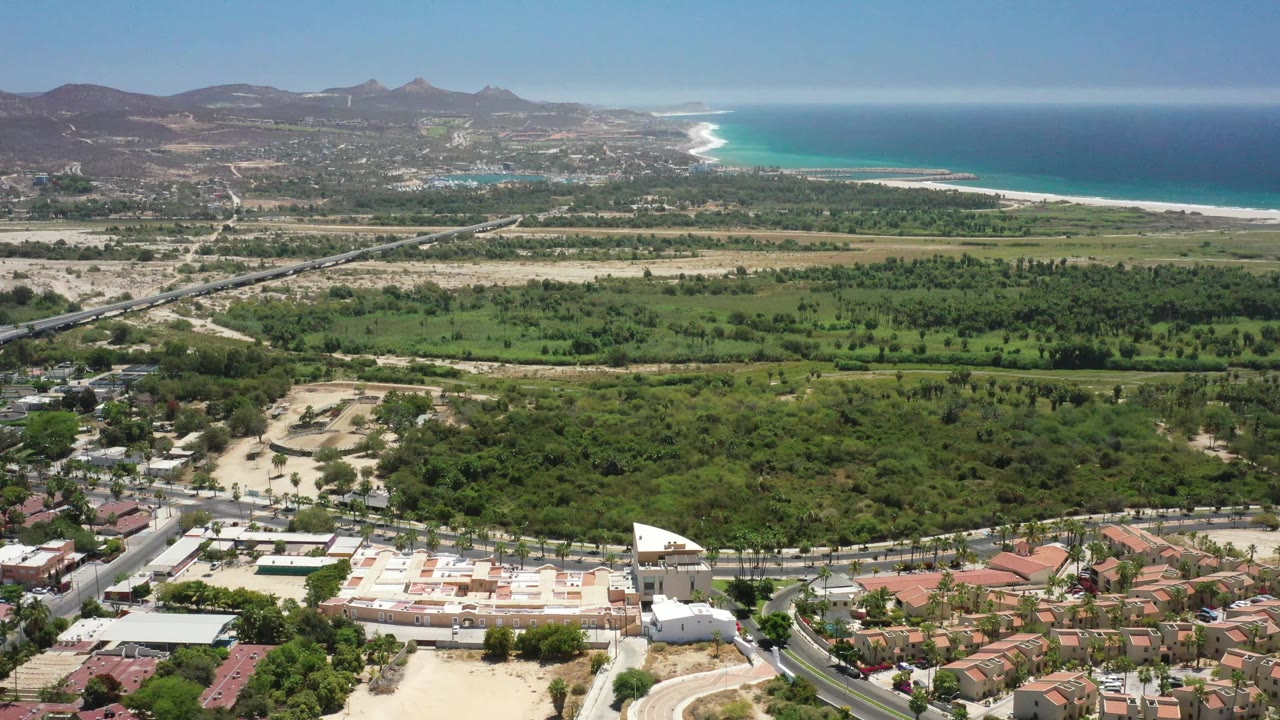 San Jose del Cabo aerial frame showing neighborhood roofs along the bottom, a road crossing the green midground, arid hills in the distance, bright blue ocean to the right, and no visible people.