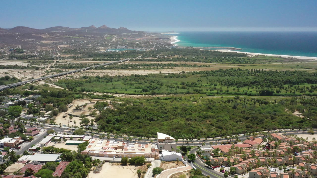 Drone view across green lowlands toward hills and a bright ocean shoreline.