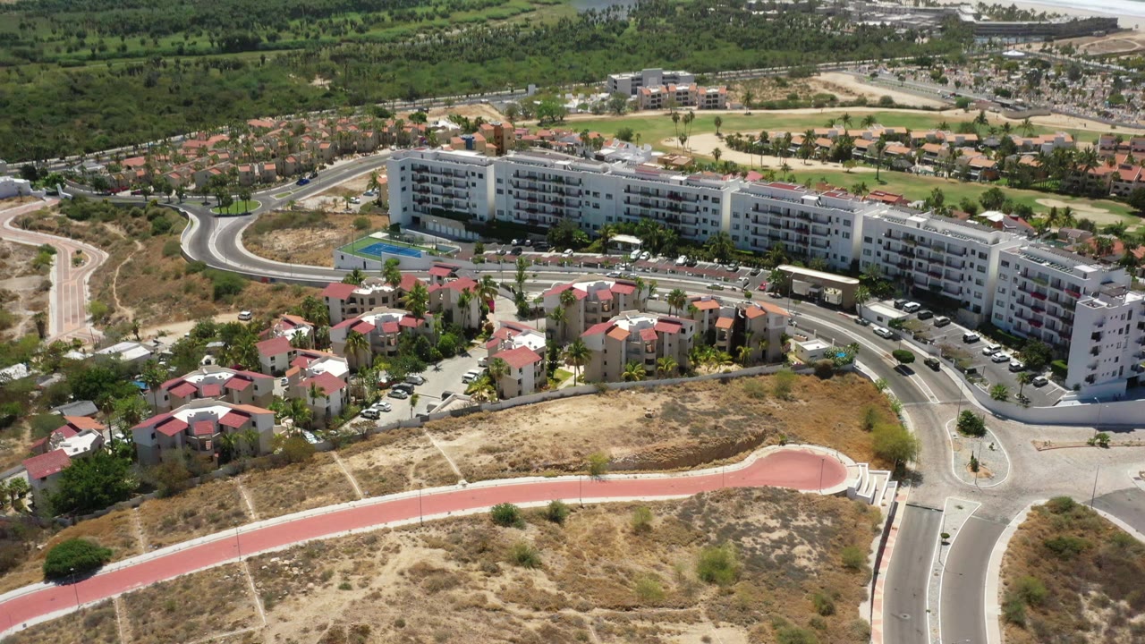 High aerial frame in San Jose del Cabo showing the long white apartment block slightly more centered, small homes below it, curving roads, green inland vegetation, beach, and blue ocean beyond, with no visible people.