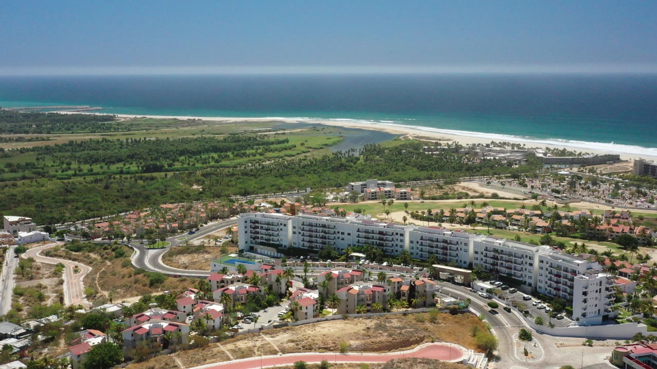 Opening aerial frame over San Jose del Cabo, Mexico, with white beachfront buildings, red-roofed homes, curving roads, green coastal land, pale beach, blue ocean, and no visible people.