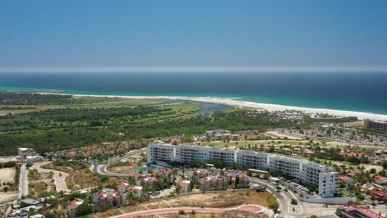 Wide aerial view over San Jose del Cabo with white mid-rise buildings, red-roofed homes, curving roads, green coastal land, beach, ocean horizon, and no visible people.