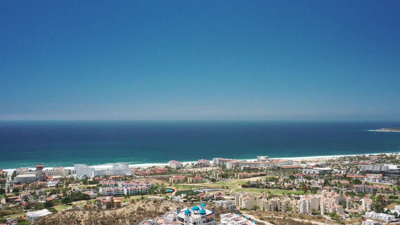 Opening aerial frame over San Jose del Cabo, Mexico, with low-rise buildings, roads, sandy beach, turquoise water, dark blue sea, clear sky, and no visible people.