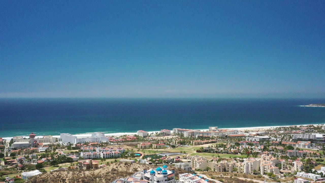High view over San Jose del Cabo with beachfront buildings, inland roads, a pale beach strip, layered blue water, distant offshore land, and no visible people.