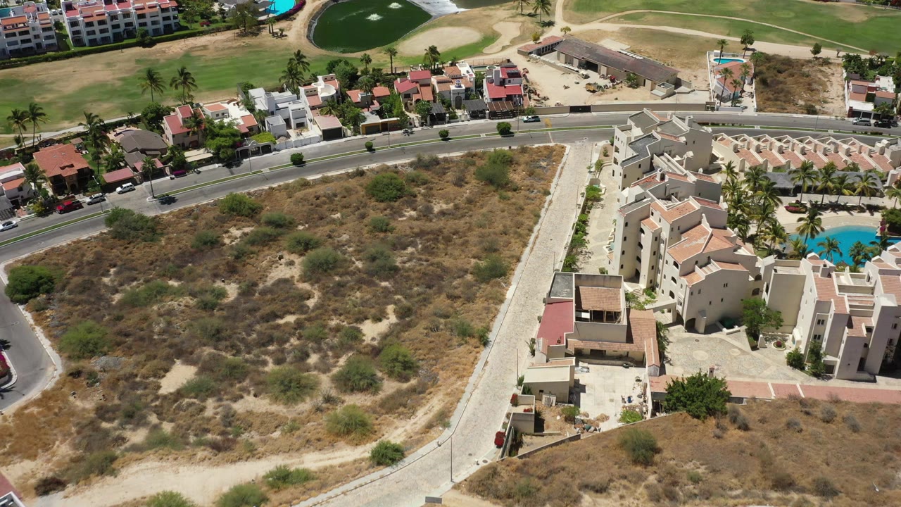 High overhead view in San Jose del Cabo with tan villas at right, open scrubland in the center, a curved road near the top, golf turf, and bright pool water, with no visible people.