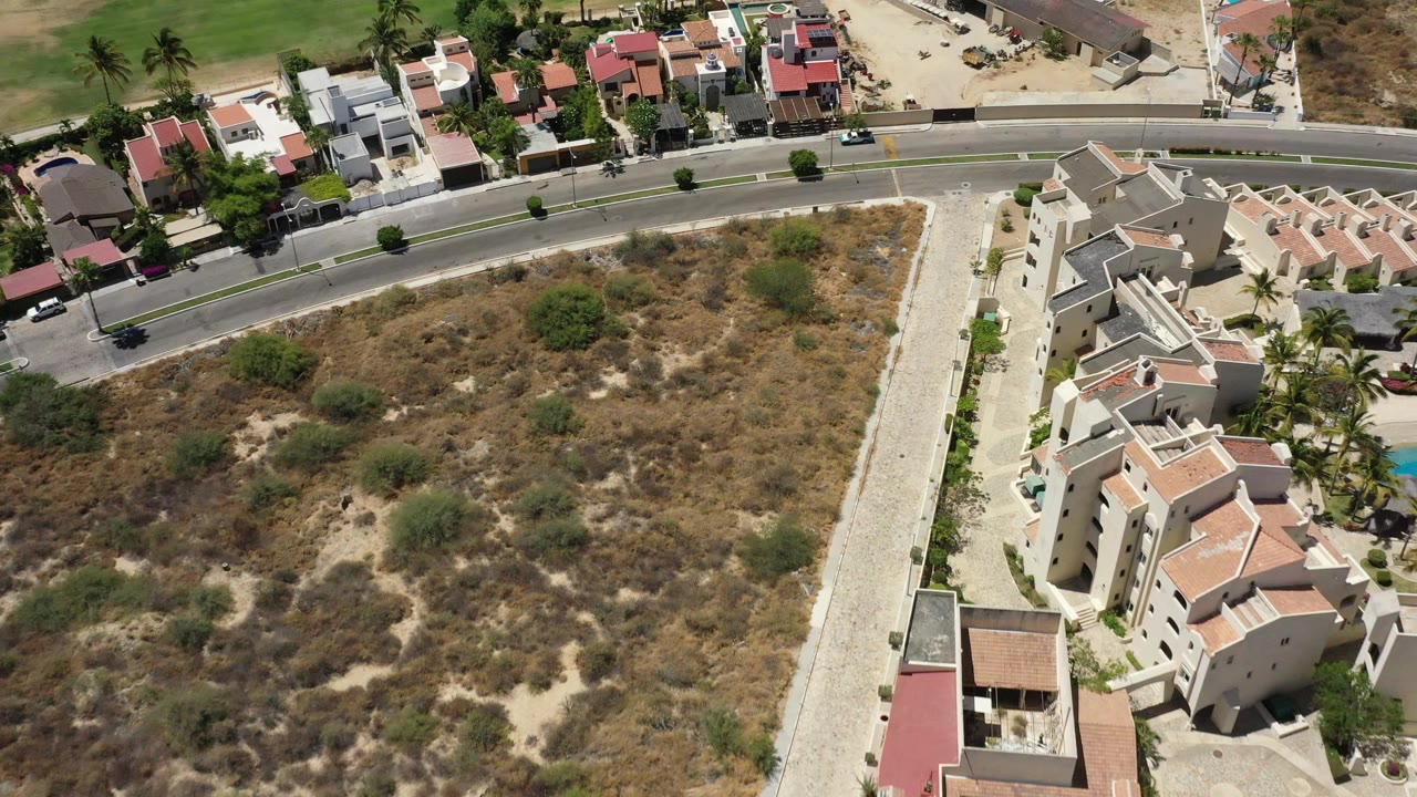 At 0:01, the San Jose Del Cabo aerial view centers on a dry open parcel, with the road above it and residential buildings running down the right side, with no visible people.