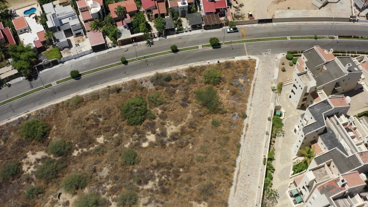 Aerial San Jose Del Cabo frame with a broad dry lot below a curved residential street, private homes and pools along the road, and no visible people.
