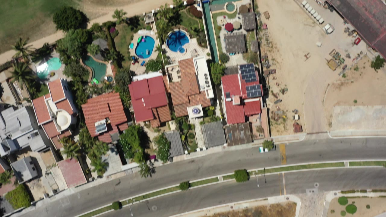 Straight-down aerial frame over San Jose del Cabo homes with red roofs, blue pools, palm trees, a curved paved road, sandy ground, and no visible people.