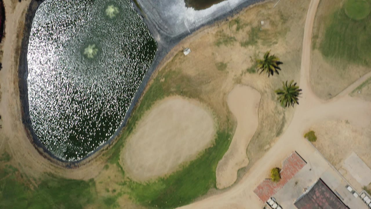 Overhead view of a small lake, sand area, pathways, palms, and golf-course grass.
