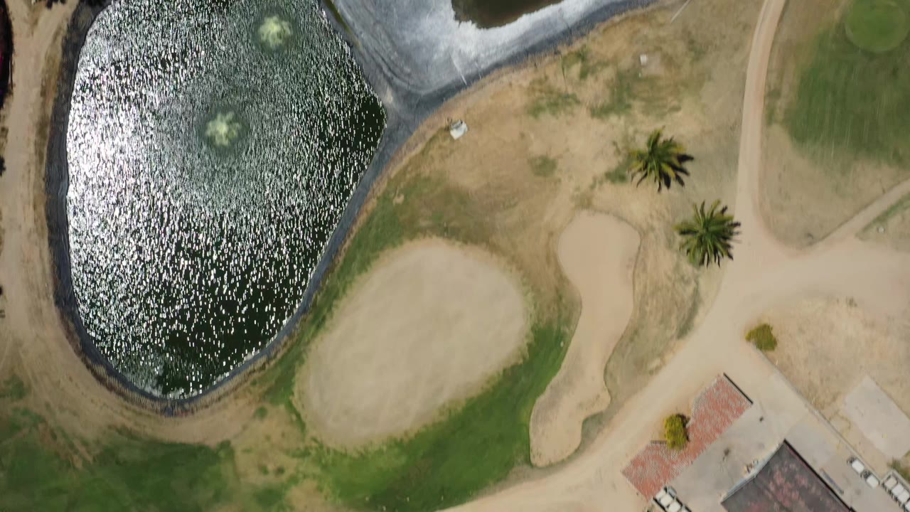 Aerial San Jose del Cabo frame with the lake shifted left in view, a rounded sand feature below it, curved pathways, palms, and no visible people.